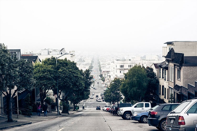 photograph looking downhill over a neighborhood street, homes lining the side, cars parked on the right