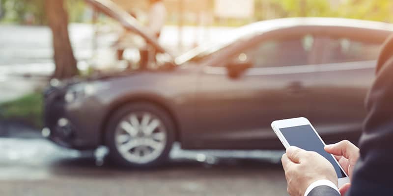 businessman using his cell phone to call for roadside assistance; his car in the background visible, but out of focus