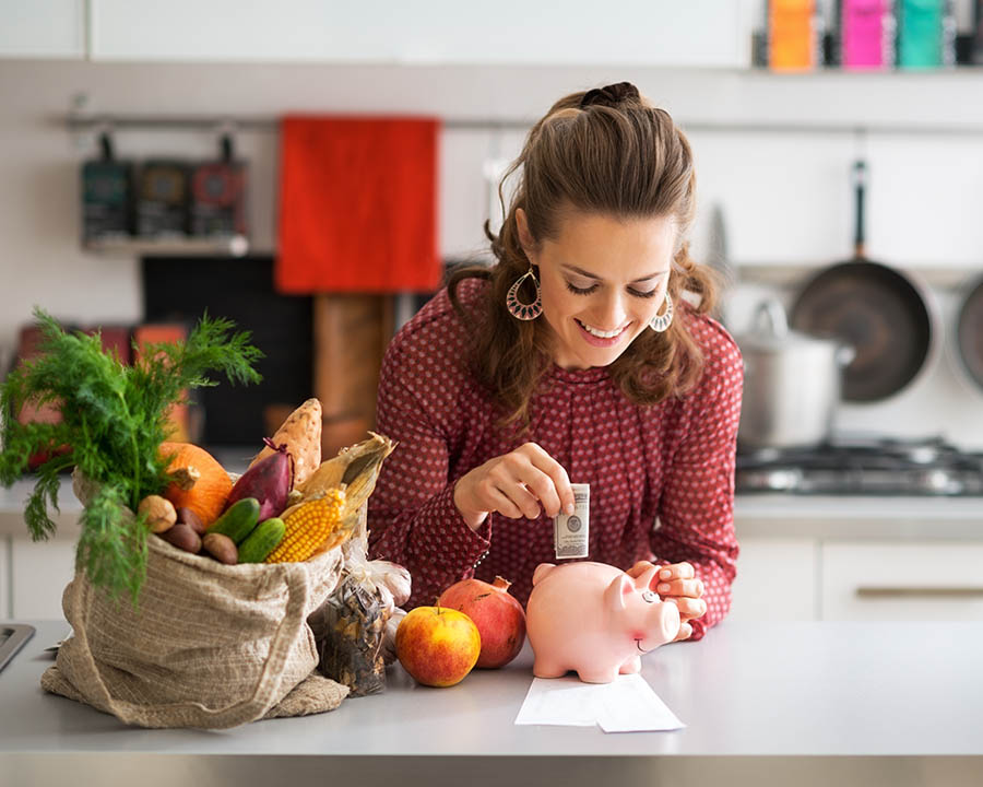 Picture of a woman in a kitchen with food, placing dollar bills into a piggy bank.